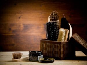 A wicker basket holds various brushes and polishing tools against a wooden background. In front, an open black container with cream and a small brush are placed on a wooden surface, perfect for understanding shoe brushes and choosing the right shoe brush for your needs.