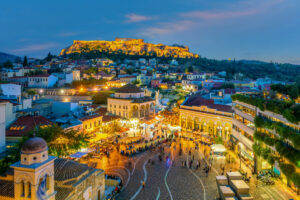 Aerial view of a bustling city square in Athens at dusk, one of Europe's prime holiday destinations. Crowds gather below, surrounded by vibrant buildings. In the background, the Acropolis is illuminated atop a hill, lively with warm lights contrasting against the deepening blue evening sky.